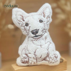 Könnte beinhalten: Ein weißes Plüschtier in Form eines Bullterriers mit braunen Stickereien, die die Gesichtszüge, Ohren und den Körper betonen. Das Wort naduwu ist in der oberen linken Ecke in einem dunkelbraunen ovalen Logo aufgedruckt.