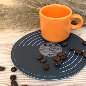 May include: A gray vinyl record-shaped coaster with white stitching and the text "Side A" in the center. An orange ceramic mug sits on top of the coaster, and coffee beans are scattered around the coaster.