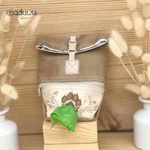 May include: A tan roll-top bag with a beige front pocket featuring an embroidered paw print design. The bag includes a plastic buckle closure, a zippered front compartment, and a green plastic bag dispensing through the embroidered paw opening.