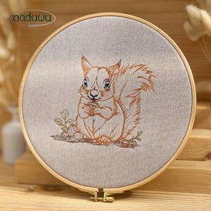May include: Embroidered artwork featuring a detailed orange squirrel holding a nut, set against a neutral fabric background. The design is framed within a wooden embroidery hoop. The brand name "naduuu" is visible in the upper left corner.