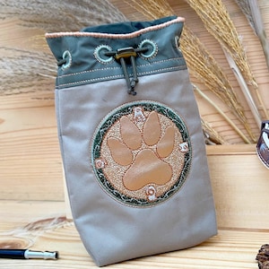Könnte beinhalten: Eine graue Kordeltasche mit grünem Oberteil und einem braunen Pfotenabdruck, der auf der Vorderseite gestickt ist. Die Tasche ist aus einem weichen, strapazierfähigen Stoff gefertigt und hat einen Kordelverschluss.