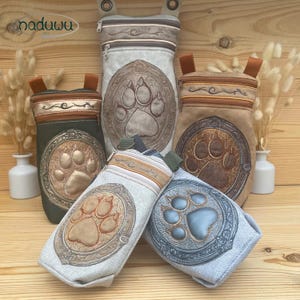 May include: A collection of zippered pouches in various sizes and colors, each featuring an embroidered paw print design within an ornate frame. The pouches are in shades of green, tan, and gray, with brown and gold accents. The brand name "naduuu art" is visible.