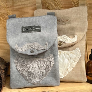 Könnte beinhalten: Zwei graue und beigefarbene Leinentaschen mit gestickten Schnurrbärten und Bärten. Die Taschen haben eine Klappe mit dem Text "Beard Care" darauf. Die Taschen liegen auf einer Holzoberfläche.