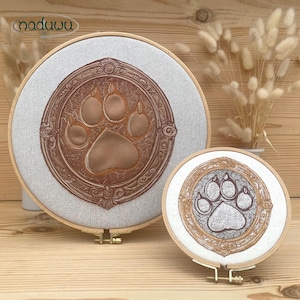 Pode incluir: Dois bastidores bordados com um desenho de pegada. O bastidor maior apresenta uma pegada castanha com uma borda decorativa castanha, sobre um tecido cinza claro. O bastidor menor tem um design semelhante. Ambos estão em bastidores de madeira.