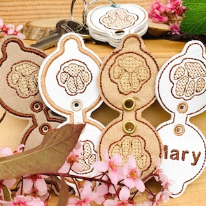 Può includere: Quattro portachiavi con un design a forma di impronta di zampa. Due sono beige con cuciture marroni e due sono bianchi con cuciture marroni. Un portachiavi bianco ha il nome "Mary" ricamato.