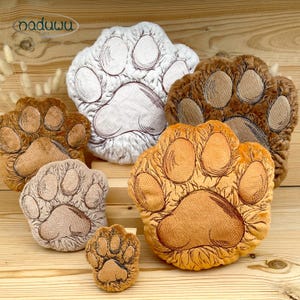 May include: A collection of five plush paw-shaped pillows in varying sizes and colors, including white, light brown, medium brown, and dark brown. Each soft pillow features embroidered details outlining the paw pads and toes in a dark thread.