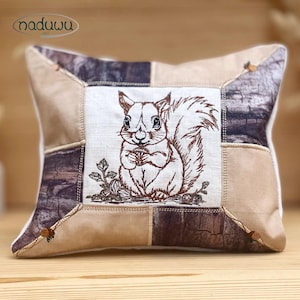 May include: Square cushion with a patchwork design. The centre features an embroidered squirrel holding a nut. The cushion has a neutral colour palette with shades of brown, beige, and cream. Small wooden beads adorn the corners.