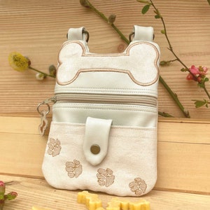 Könnte beinhalten: Eine weiße Leder-Hunde-Leckerli-Tasche mit einem Knochen-Reißverschluss-Zipper und Pfoten-Stickerei. Die Tasche hat eine kleine Tasche auf der Vorderseite und ein Reißverschlussfach auf der Rückseite.