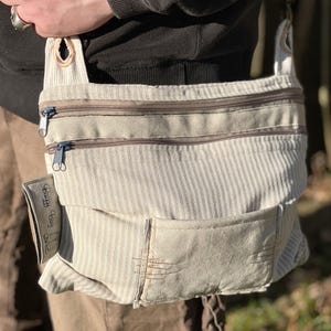 Puede incluir: Un bolso bandolera de lona a rayas blancas y marrones con un bolsillo con cremallera y un pequeño parche que dice "Earth Day Bag".