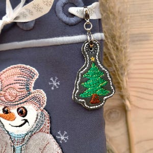 May include: A grey fabric bag with a snowman and a Christmas tree charm. The snowman is wearing a pink hat and a brown coat. The Christmas tree charm is green with a gold star on top.