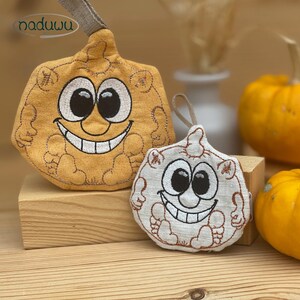 May include: Two embroidered pumpkin-shaped ornaments with smiling faces. One is a light orange colour, and the other is white. Both have black eyes and a wide, happy grin. They have a loop at the top for hanging. The ornaments are on a wooden block.