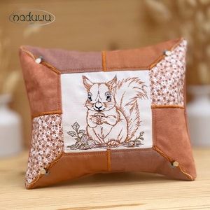 May include: Square cushion with a brown suede-like exterior and a central embroidered squirrel design. The squirrel is holding a nut, with floral accents. The cushion has a patchwork design with floral and solid brown panels. The brand name "naduwu art" is in the upper left corner.