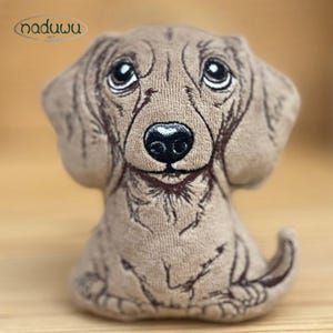 Puede incluir: Un juguete de peluche de perro salchicha sentado en tonos marrones. El juguete tiene ojos grandes y expresivos, una nariz negra y detalles bordados. La marca "naduwu art" está en un logotipo circular en la esquina superior izquierda.