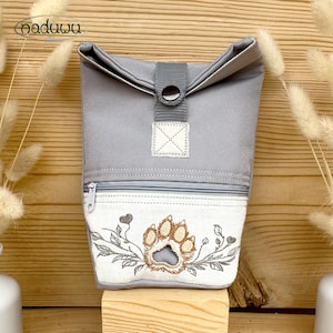 May include: A grey roll-top bag featuring a central snap closure and a white front pocket with a zipper. The pocket displays an embroidered brown paw print surrounded by grey leafy vines and small heart shapes. The bag has a structured fabric design.