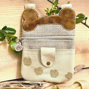 May include: A beige and brown fabric dog bone-shaped zippered pouch with a paw print design. The pouch has a zippered pocket on the front and a small zippered pocket on the back.