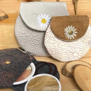 Puede incluir: Tres pequeñas bolsas redondas de tela con diseños de margaritas bordadas. Las bolsas están hechas de diferentes telas, incluyendo denim, lino y arpillera. Las bolsas se cierran con una solapa y un botón.