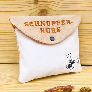May include: A beige and white pouch with the text "SCHNUPPER-KURS" embroidered in orange. The pouch has a flap secured by a black snap and a cartoon dog face on the front. The pouch is on a wooden surface.