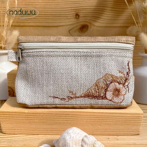 May include: A small, rectangular pouch with a zipper closure. The pouch is made of a light gray woven fabric with a brown embroidered floral design. The top and bottom edges are trimmed with a tan fabric. The brand name "naduuu" is printed on the top left.