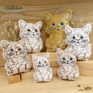 May include: A collection of five plush cat toys featuring embroidered details. The set includes one golden-yellow cat and four white cats with brown tabby-style markings. Each cat has large blue eyes, whiskers, and a small nose, presented as soft figures.