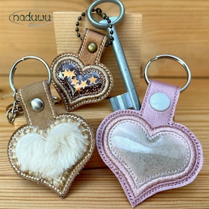 May include: Three heart-shaped keychains in various designs. One has a fluffy white center, another features gold stars, and the third is a clear, pink-stitched design. Each keychain has a metal ring and a leather strap.
