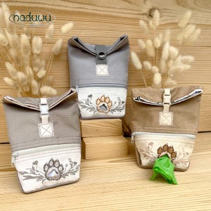 May include: Three roll-top pouches in gray and tan canvas, each featuring a front zippered pocket with embroidered paw prints and floral motifs. The pouches have plastic buckle closures and one displays a green plastic bag dispensing from the front.