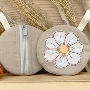 May include: Two beige round pouches with white fabric loops. One pouch has a white daisy embroidered on it with a yellow center. The other pouch has a silver zipper. The pouches are made of a soft fabric.