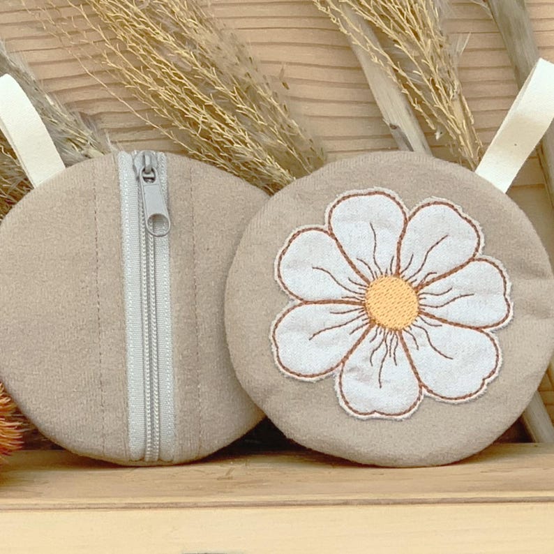 May include: Two beige round pouches with white fabric loops. One pouch has a white daisy embroidered on it with a yellow center. The other pouch has a silver zipper. The pouches are made of a soft fabric.