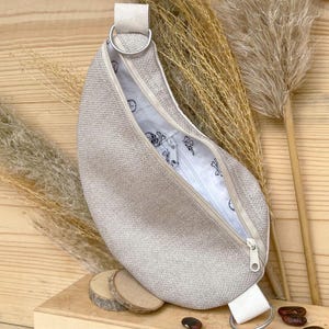 Puede incluir: Una bolsa de color gris claro en forma de media luna con cierre de cremallera y una correa blanca. La bolsa está hecha de un tejido texturizado y tiene un forro blanco con un pequeño diseño negro.