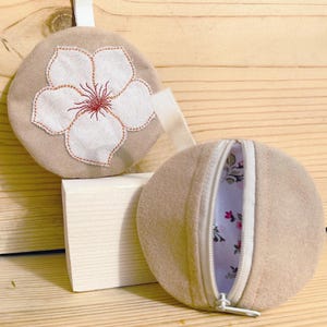May include: Two beige, round pouches. One features a white flower with brown stitching. The other is open, revealing a floral patterned interior. Both have a white zipper and a small loop.