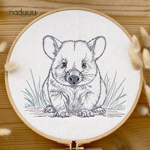 Könnte beinhalten: Ein runder hölzerner Stickrahmen mit weißem Stoff, der eine detaillierte schwarze Strichzeichnung eines Wombats zeigt. Der Wombat hat braune Augen und ist von dünnen grünen Grashalmen umgeben. Das Logo naduwu ist in der oberen linken Ecke aufgedruckt.