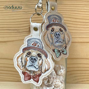 Peut inclure: Deux porte-clés brodés représentant un Golden Retriever portant un chapeau haut de forme et un nœud papillon. Les porte-clés sont beiges et crème, avec des coutures détaillées en marron, beige et bleu. Attaches en métal argenté.