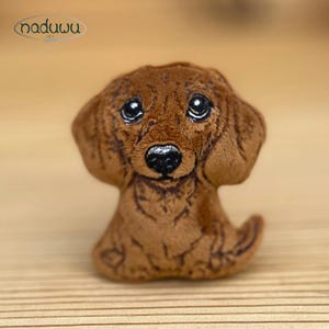 May include: A brown plush dachshund dog figurine with large, expressive eyes and a black nose. The dog is sitting, with its tail curled up. The figurine has a soft, textured appearance and is likely handmade. The logo "naduuu art" is in the upper left corner.