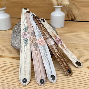 以下が含まれることがあります： 様々な色の刺繍入り花柄デザインが施された、長く細い布製リストバンド4本セット。各バンドの両端には金属製のスナップボタンが付いています。色はクリーム色、薄いピンク色、薄い灰色、濃い茶色が含まれています。