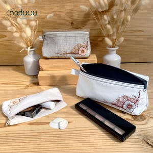 May include: A collection of cosmetic bags in various sizes and materials. The bags are white, beige, and gray, with a seashell and floral embroidery design. One bag is open, revealing makeup products. The bags are displayed on a wooden surface.