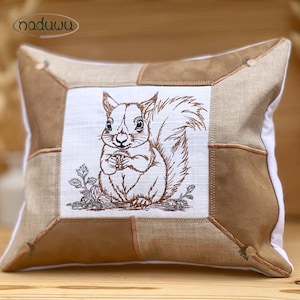 Könnte beinhalten: Quadratisches Zierkissen mit einem gestickten Eichhörnchen-Design. Das Kissen hat eine weiße Mitte mit einer braunen Eichhörnchen-Stickerei, umgeben von einem Patchwork aus beigen und braunen Stoffen. Der Markenname "naduuu" befindet sich in der oberen linken Ecke.