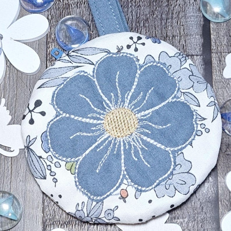 May include: A round, floral-patterned coaster with a blue flower design and a light blue loop. The coaster features a white background with blue floral accents and a gold center. Decorative elements include white paper cutouts and glass marbles.