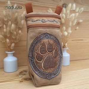 Könnte beinhalten: Eine beigefarbene Tier-Tragetasche mit einem großen, gestickten Pfotenabdruck-Design. Die Tasche hat einen Reißverschluss oben und braune Träger. Das Design umfasst einen dekorativen Rahmen und den Markennamen "naduuu".