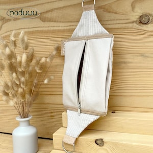 Può includere: Una pochette color crema con cerniera, un accento in tessuto a righe e un anello di metallo. La pochette è appesa su uno sfondo di legno. Il marchio "naduuu" è visibile nell'angolo in alto a sinistra.