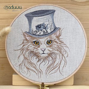 Könnte beinhalten: Gesticktes Kunstwerk mit einer Katze, die einen Zylinder mit einem Fisch und Wasserpflanzen trägt. Die Katze hat gelbe Augen und braunes Fell. Die Stickerei befindet sich in einem runden Holzrahmen. Das Kunstwerk ist mit "naduuu art" signiert.