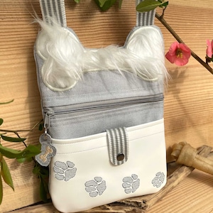 May include: A gray and white fabric crossbody bag with a furry dog bone design. The bag has a zippered pocket and a flap with a snap closure. The flap has paw prints embroidered on it.
