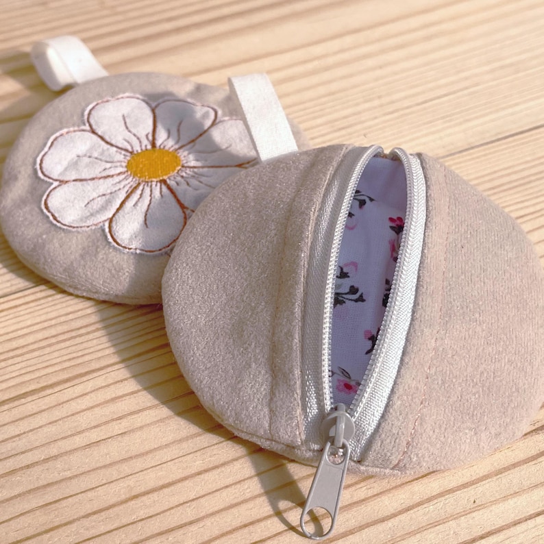 May include: Two beige round coin purses. One features a white daisy embroidery with a yellow center. The other is partially unzipped, revealing a floral patterned interior. Both have a silver zipper and a small fabric loop.