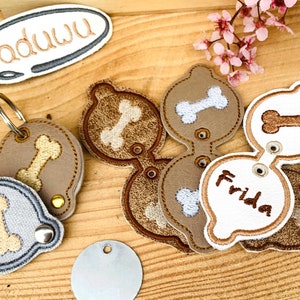 以下が含まれることがあります： 骨のデザインが施された犬をテーマにしたキーホルダー5個セット。キーホルダーは茶色と灰色の生地で作られており、白い骨のデザインが施されています。1つのキーホルダーには「Frida」という名前が刺繍されています。