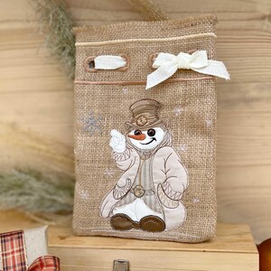 Puede incluir: Una bolsa de regalo de arpillera con un diseño de muñeco de nieve. El muñeco de nieve lleva un sombrero de copa marrón, un abrigo marrón y botas marrones. La bolsa tiene una cinta blanca atada alrededor de la parte superior.