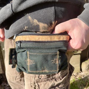 May include: A dark green canvas fanny pack with a cork top zipper and a small pocket on the front. The fanny pack has a brown leather strap with the text "Belly Bag Co." printed on it.