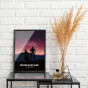 May include: A black framed poster with a purple and pink gradient background. Two silhouettes of figures stand in front of a large moon. The text reads "Rebellions are built on hope. Cassian Andor."