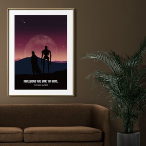 May include: A minimalist poster with a dark purple and pink gradient background featuring two silhouettes of figures standing on a hilltop under a large moon. The text "Rebellions are built on hope." is displayed below the figures, with the words "Cassian Andor" in smaller font below the text.