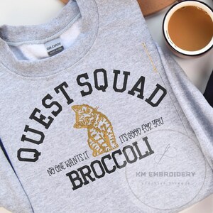 May include: Gray sweatshirt with black text that reads "Quest Squad" and "No one wants it, it's good for you Broccoli". A gold cat illustration is in the center of the text.