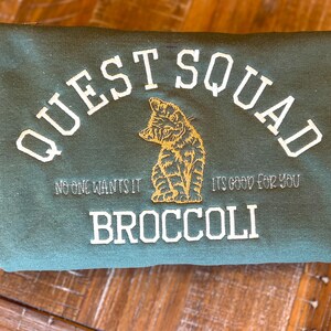 May include: A dark green sweatshirt with the text "QUEST SQUAD" in white embroidery. The text "NO ONE WANTS IT" and "IT'S GOOD FOR YOU" are also embroidered in white. A small embroidered image of a cat is in the center of the sweatshirt. The text "BROCCOLI" is embroidered in white below the cat.