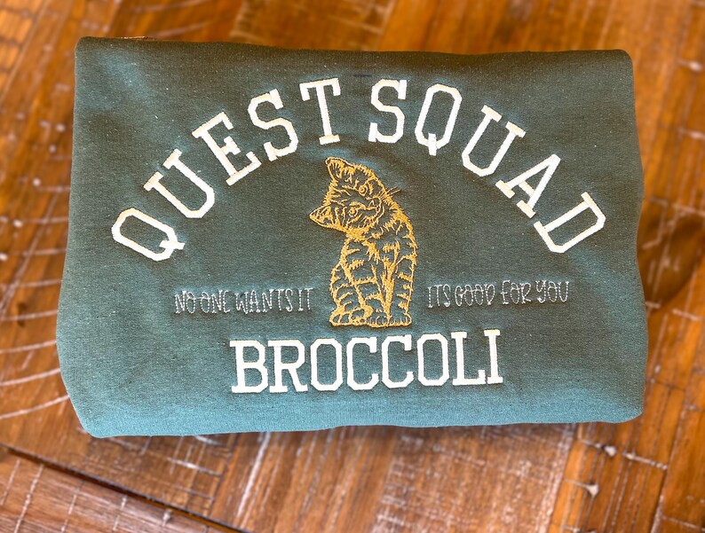 May include: A dark green sweatshirt with the text "QUEST SQUAD" in white embroidery. The text "NO ONE WANTS IT" and "IT'S GOOD FOR YOU" are also embroidered in white. A small embroidered image of a cat is in the center of the sweatshirt. The text "BROCCOLI" is embroidered in white below the cat.