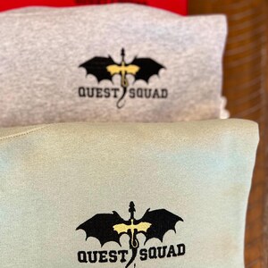 May include: Three sweatshirts stacked on top of each other. The top sweatshirt is red, the middle sweatshirt is gray, and the bottom sweatshirt is light green. Each sweatshirt has a black dragon with yellow wings and the text "QUEST SQUAD" embroidered on the front.